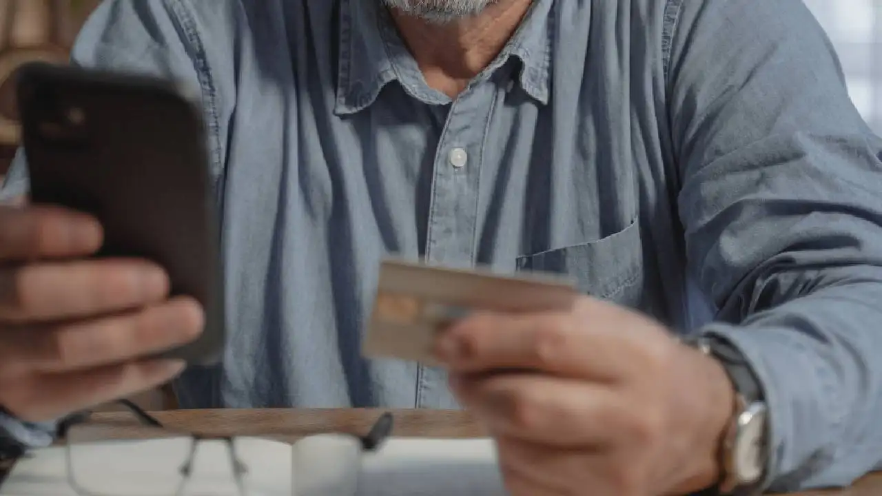 Un hombre mayor con el móvil en una mano y una tarjeta de crédito en la otra