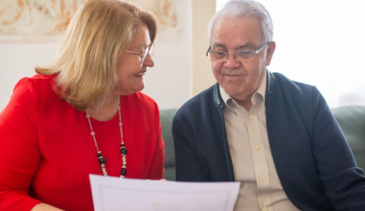 Pareja de ancianos consultando opciones de pensiones, sonriendo en un sof&aacute;, discutiendo documentos.