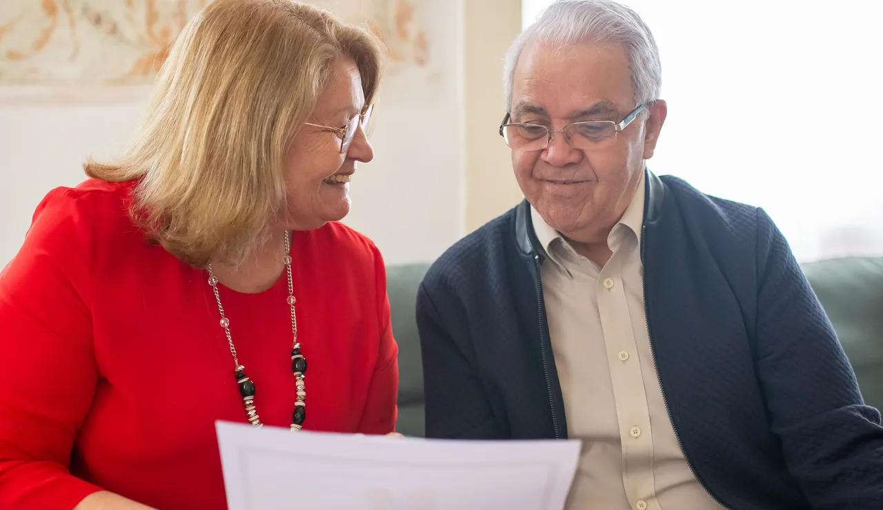 Pareja de ancianos consultando opciones de pensiones, sonriendo en un sofá, discutiendo documentos.