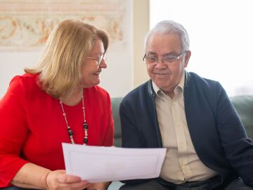 Pareja de ancianos consultando opciones de pensiones, sonriendo en un sof&aacute;, discutiendo documentos.