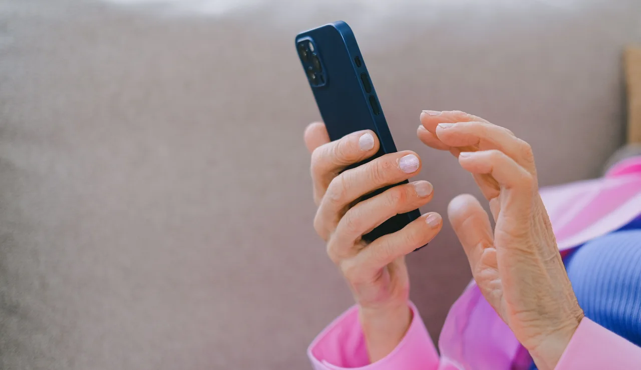 Un adulto mayor con chaqueta rosa usando un smartphone mientras se relaja en un sofá en interiores
