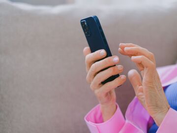 Un adulto mayor con chaqueta rosa usando un smartphone mientras se relaja en un sof&aacute; en interiores