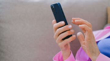 Un adulto mayor con chaqueta rosa usando un smartphone mientras se relaja en un sof&aacute; en interiores