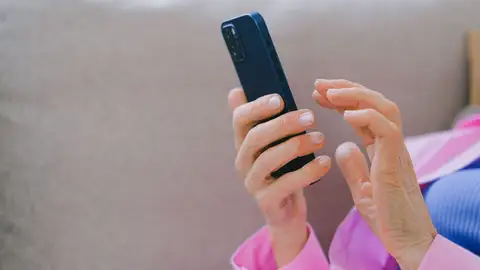 Un adulto mayor con chaqueta rosa usando un smartphone mientras se relaja en un sofá en interiores Un adulto mayor con chaqueta rosa usando un smartphone mientras se relaja en un sofá en interiores