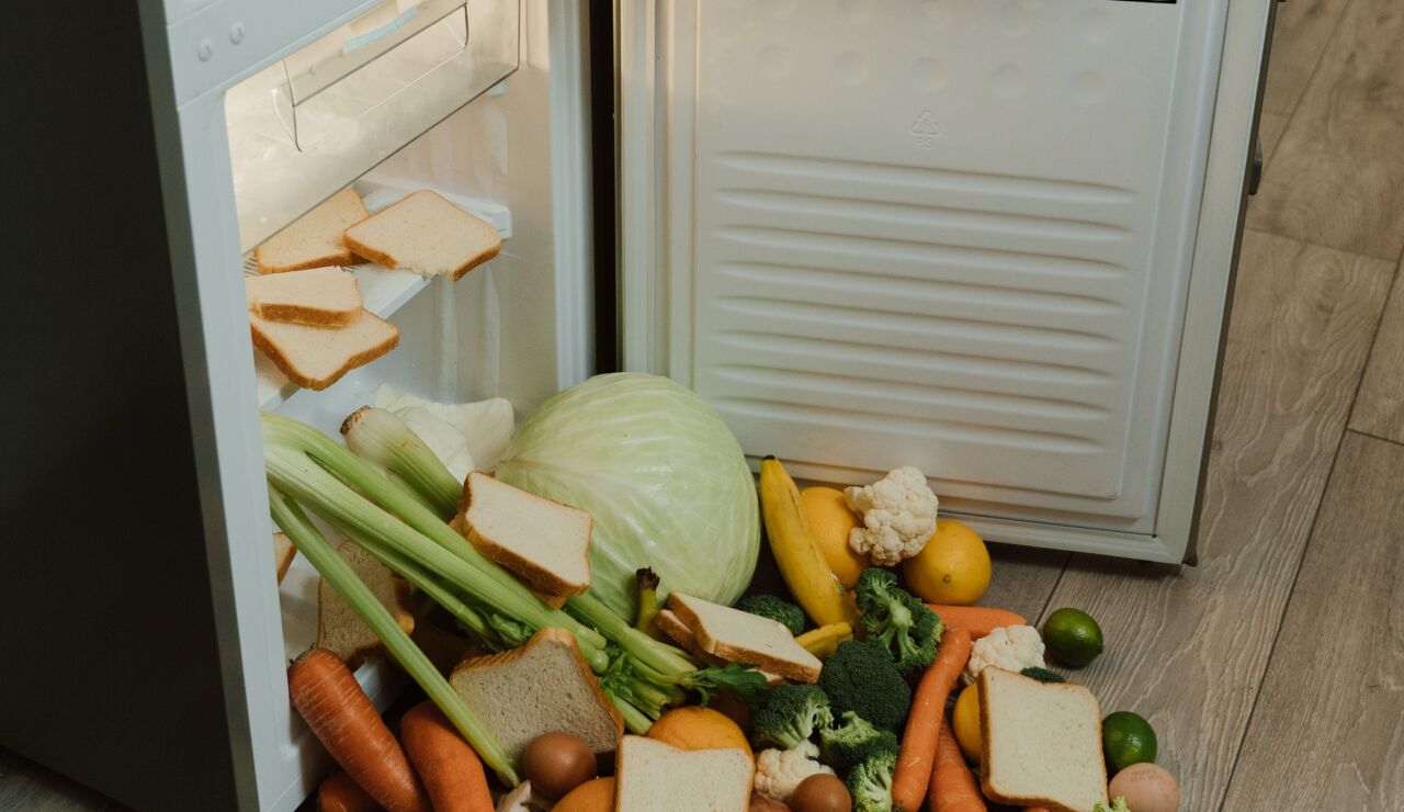 Una nevera desordenada y abierta con verduras frescas y pan esparcidos por el suelo