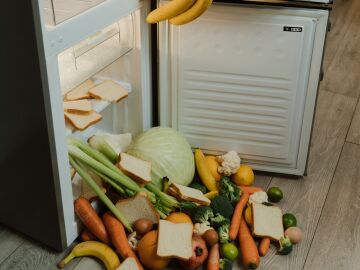 Una nevera desordenada y abierta con verduras frescas y pan esparcidos por el suelo