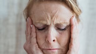 Una mujer anciana con los ojos cerrados, toc&aacute;ndose la cara, mostrando signos de estr&eacute;s y preocupaci&oacute;n en interiores.