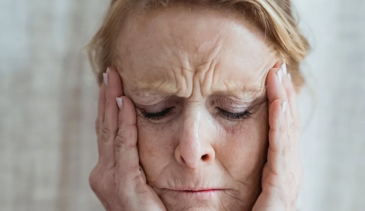 Una mujer anciana con los ojos cerrados, tocándose la cara, mostrando signos de estrés y preocupación en interiores.
