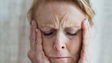 Una mujer anciana con los ojos cerrados, toc&aacute;ndose la cara, mostrando signos de estr&eacute;s y preocupaci&oacute;n en interiores.
