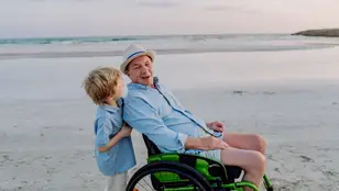 Hombre mayor en silla de ruedas empujado por un niño en la playa