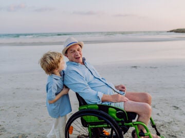 Hombre mayor en silla de ruedas empujado por un niño en la playa