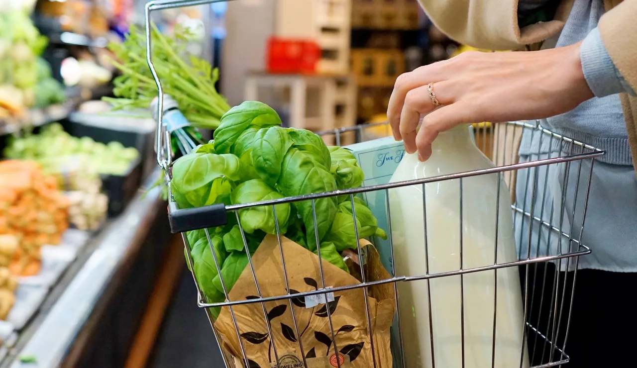 Una persona compra en un supermercado con una cesta que contiene verduras frescas, leche y hierbas.