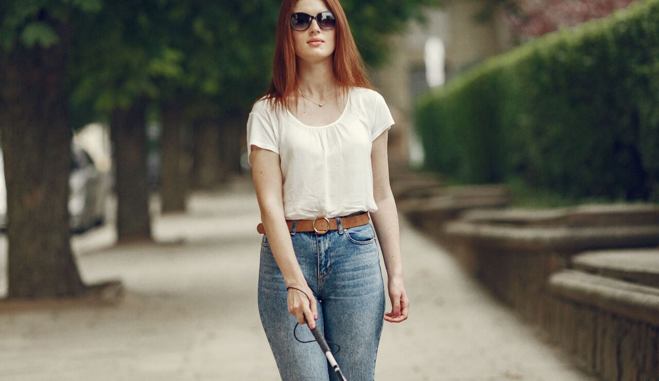 Una mujer con discapacidad visual caminando con confianza por un camino arbolado con su bast&oacute;n blanco.